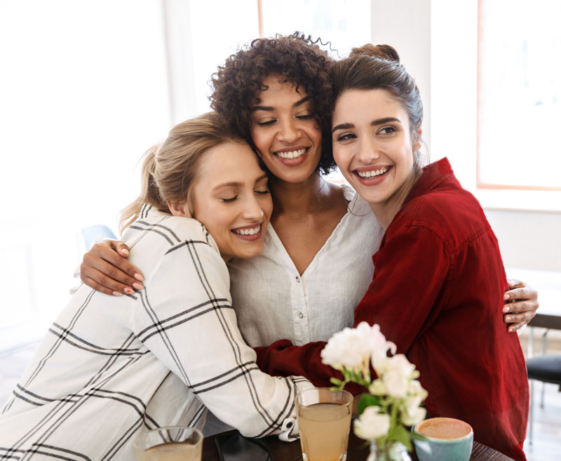 Three women are hugging at a table with flowers.